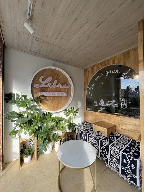 Góc trong nhà với biển neon Libra Kitchen, cây xanh và dòng chữ How was your day?